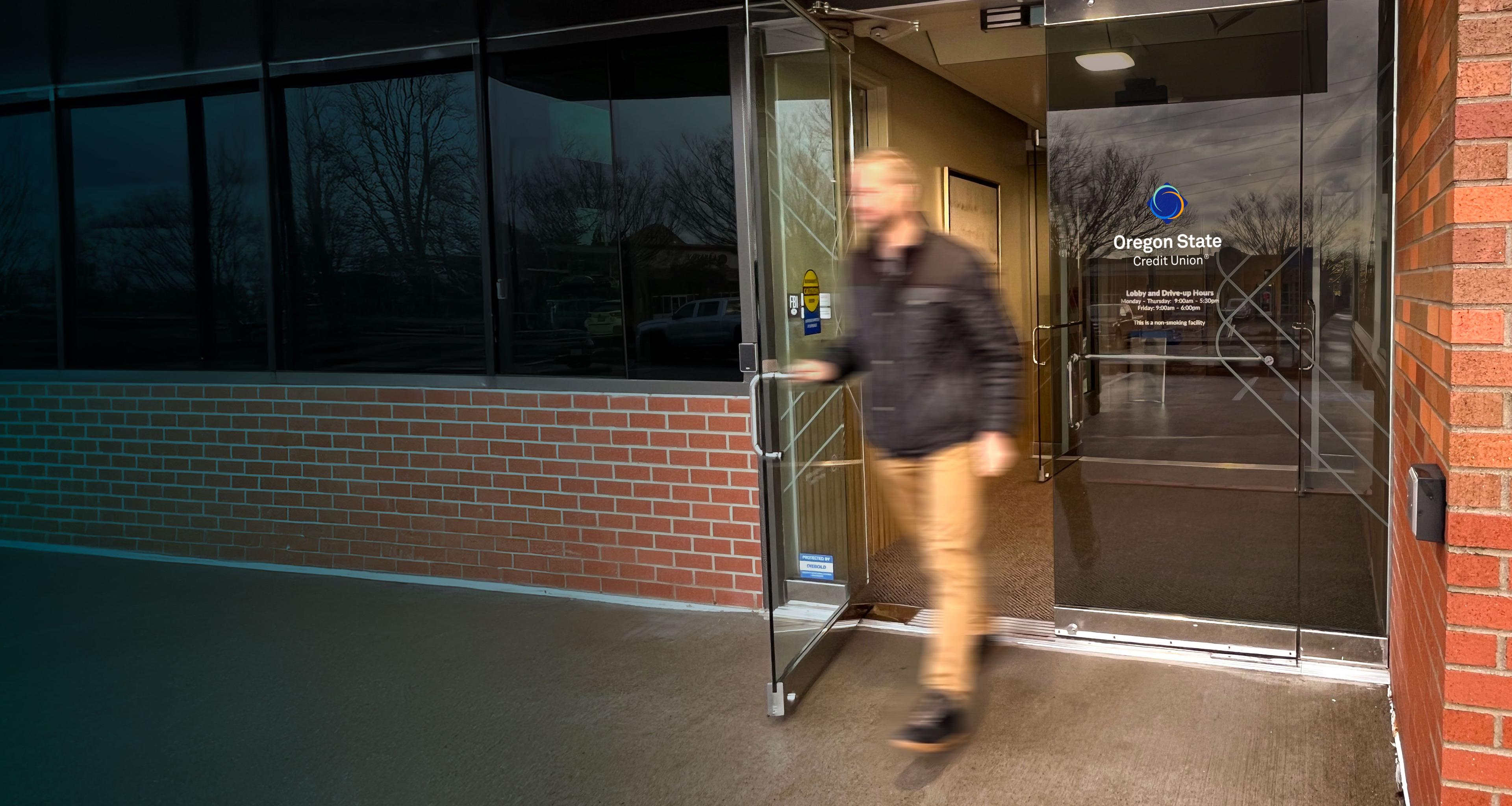 A man in a dark jacket and tan pants walking out through the glass entrance doors of an Oregon State Credit Union branch, with a brick exterior and the credit union’s logo visible on the glass behind him.